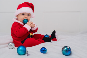 Fototapeta na wymiar Baby boy dressed as Santa is playing with Christmas decorations on the bed.