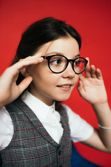 preteen girl adjusting eyeglasses and smiling while looking away isolated on red