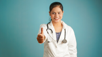 Portrait of a young female doctor gesturing thumbs up, Cheerful Asian Indian woman doctor in apron and stethoscope isolated over blue studio background