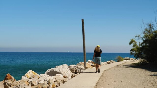 A Girl In Shorts, Jersey And Baseball Cap Walks Along Promenade Along Sea. She Has An Action Camera In Her Hand. In Background Is A Beautiful Sea And A Ship. Back View. Woman Blogger A Seaside Resort