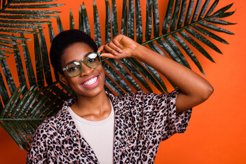 Portrait of attractive cheerful trendy girl resting in tropics touching specs isolated over bright orange color background