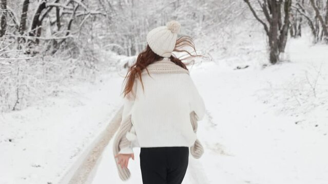 Rear view, camera follows a young long-haired woman girl running through the snowy forest of the park in a white sweater, hat and scarf, turns to the camera, smiles and extends her hand
