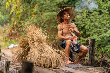Photo of an asian old male farmer village man wearing conical hat taking a break while delivering...