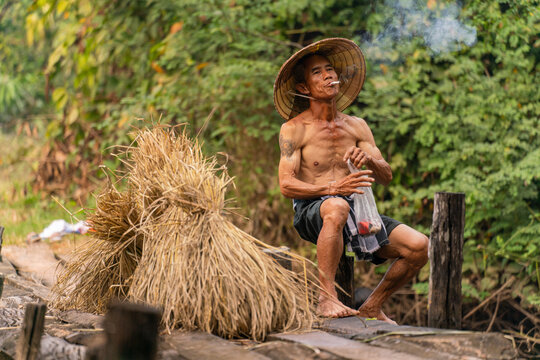 Photo of an asian old male farmer village man wearing conical hat taking a break while delivering some dry grasses for animal feeds rest sitting on a wooden bridge smoke a local tobacco