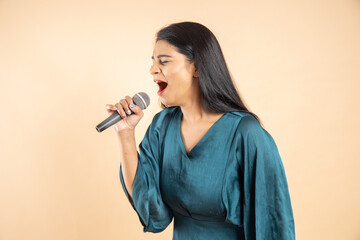 Beautiful young indian girl singer holding mic singing song isolated over beige studio background.