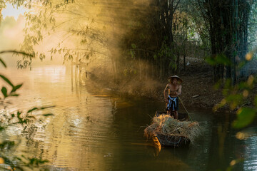 Photo of a local asian old man male boatman wearing conical hat rowing a small wooden boat across a small river during sunset time in a bamboo forest to deliver some dry grasses as animal feeds. 