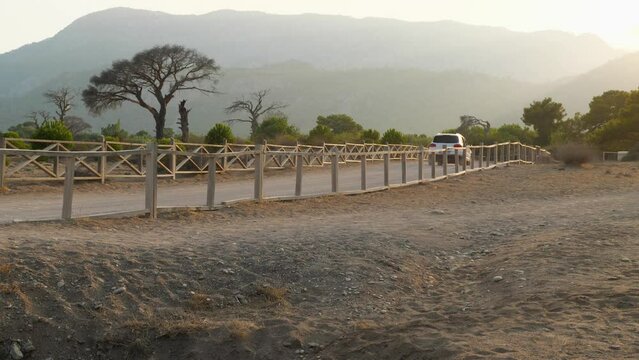 White car drives on a sandy road fenced with a wooden fence. In the background, dry trees and mountains in the rays of the setting sun. Side view. Turkish savannah in the evening on the Lycian Trail