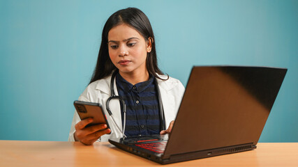 Portrait of a young female doctor working on a laptop sitting on the table or desk, Busy Asian Indian woman doctor in apron and stethoscope isolated over blue studio background, using mobile phone