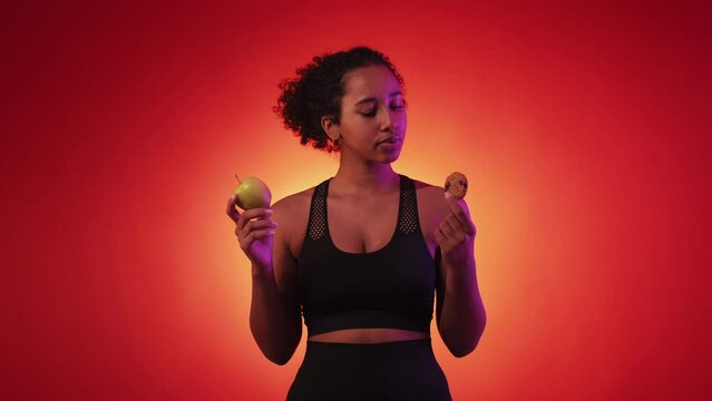 Unhealthy food. Doubtful woman. Diet problem. Sportive thoughtful lady choosing cookie in spite of apple posing neon light red yellow background.