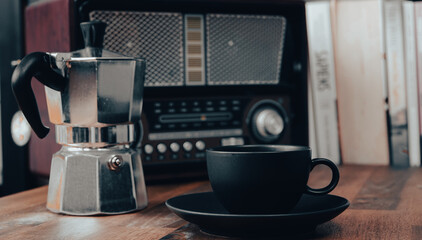 Moka pot, vintage radio, a black coffee cup and saucer on a brown wooden table, with books in the background, vintage look	