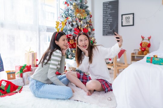 Two young asian female lady friends in a room happily celebrate Christmas together posing and gift exchange and snap photos using a vintage film camera in front a nicely decorated ornaments tree