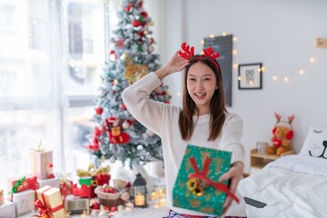 Cute beautiful young asian lady woman wearing reindeer headband holding a green color gift box with bells and posing in front a big Christmas tree with lots of decoration lights gift box and ornament