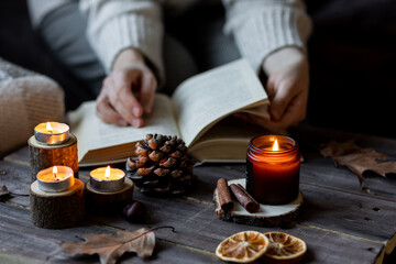 Cozy autumn or winter composition with aromatic candle, wool sweater. Aromatherapy, home atmosphere of cosiness and relax. Woman reading a book. Wooden background close up.