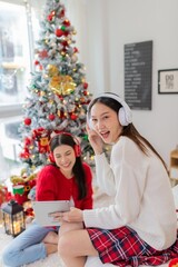 Obraz na płótnie Canvas Two young asian female lady friends in a room listening to new songs on their headphone via their digital tablet celebrate Christmas together