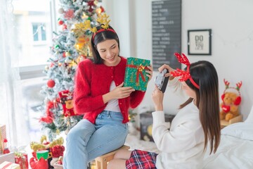 Two young asian female lady friends in a room happily celebrate Christmas together posing and gift exchange and snap photos using a vintage film camera in front a nicely decorated ornaments tree