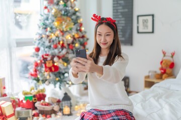 Cute beautiful young asian lady woman wearing reindeer headband selfie and video call with mobile phone posing in front a big Christmas tree with lots of decoration lights gift box and ornaments