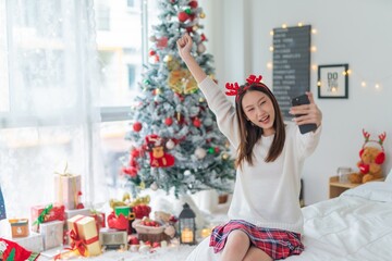 Cute beautiful young asian lady woman wearing reindeer headband selfie and video call with mobile phone posing in front a big Christmas tree with lots of decoration lights gift box and ornaments