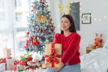 Cute beautiful young asian lady woman wearing reindeer headband holding a gold color gift box with red ribbon posing in front a big Christmas tree with lots of decoration lights gift box and ornaments