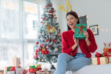 Cute beautiful young asian lady woman wearing reindeer headband holding a green color gift box with bells and posing in front a big Christmas tree with lots of decoration lights gift box and ornaments
