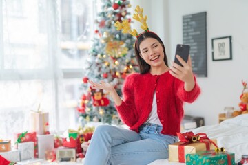 Fototapeta na wymiar Christmas celebration. Young asian lady woman wearing reindeer headband selfie and video call with mobile phone posing in front a Christmas tree with lots of decoration lights gift box and ornaments