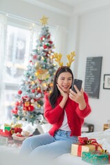 Cute beautiful young asian lady woman wearing reindeer headband selfie and video call with mobile phone posing in front a big Christmas tree with lots of decoration lights gift box and ornaments