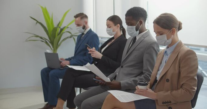 Diverse job candidates in safety mask sitting in office hallway waiting for secretary to invite them