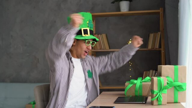 Male fan in leprechaun hat watching football play online broadcast on St.Patrick's Day on laptop, betting at bookmaker website. Lucky man celebrating victory after winning jackpot in casino.