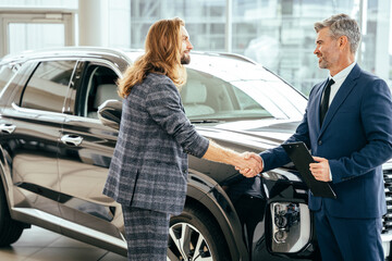 Happy middle age car salesperson came to an agreement with young long haired beard man in a showroom with new crossover on background.