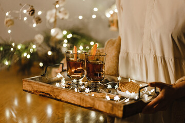 Female hands hold cup of tea next to cookies and Christmas tree on tray. Selective focus.