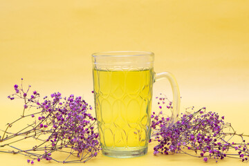 Uma caneca de vidro com chá com pequenos galhos de flores em um fundo amarelo. Formato horizontal.