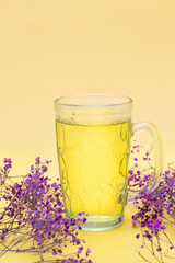 Uma caneca de vidro com chá com pequenos galhos de flores em um fundo amarelo. Formato vertical.