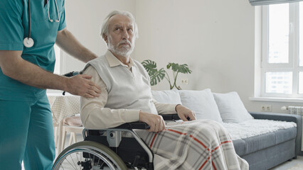 Male nurse taking care of upset senior man in wheelchair, retirement home, care