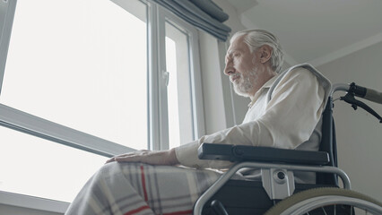 Senior man with disability feeling lonely, sitting in wheelchair near window