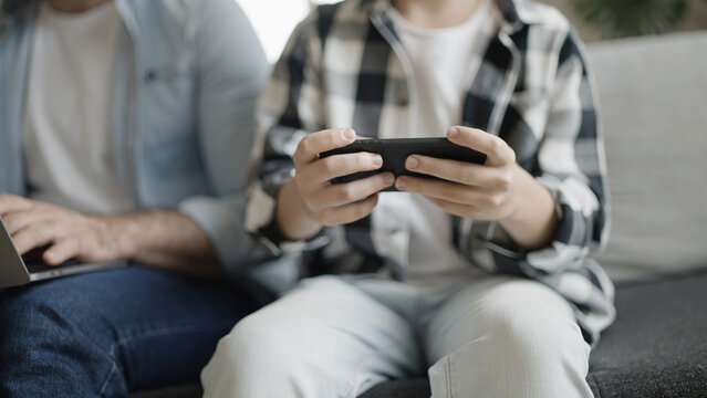 Teenage boy playing game while father typing on laptop, male pastime together