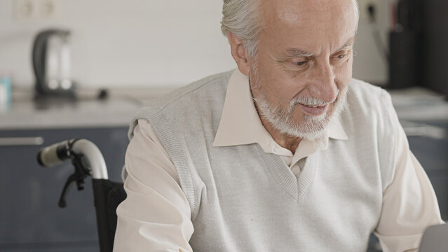 Smiling gray-haired man in wheelchair having online video call with a friend
