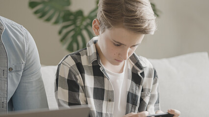 Schoolboy playing online games on smartphone while father's working on laptop