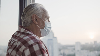 Sad old man in medical face mask looking out the window, pandemic isolation