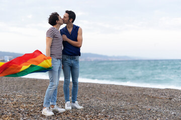  Beautiful gay young couple embraces and holds a rainbow flag. Happy couple enjoy at the beach