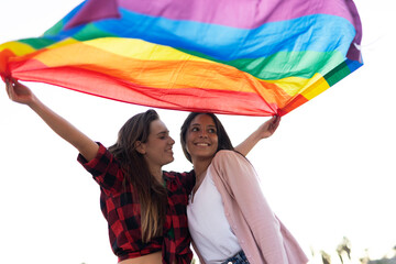 Beautiful lesbian young couple embraces and holds a rainbow flag. Girls enjoy at the beach