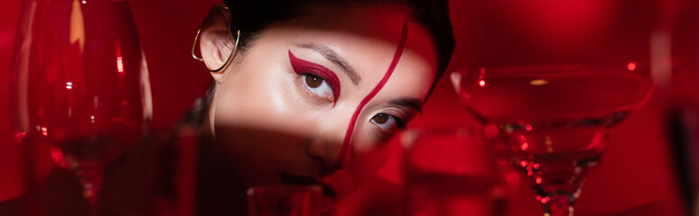 charming asian woman with bright artistic makeup looking away in light near blurred glasses on red...