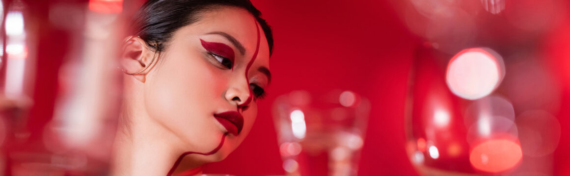 portrait of asian woman with creative makeup on face divided with line near blurred glasses on red background, banner.
