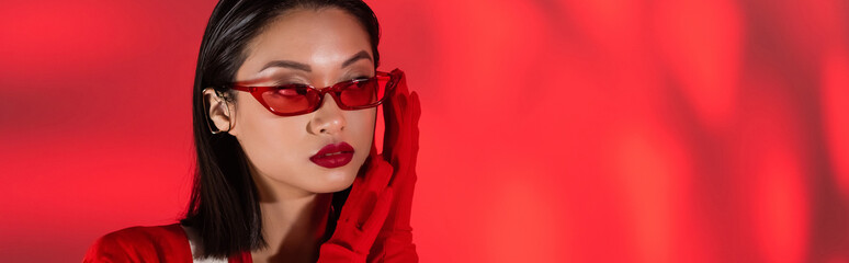 portrait of asian woman in trendy sunglasses and gloves on red abstract background, banner.