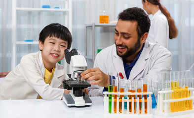 male teacher sitting at classroom desk with male student using microscope while learning science experiment