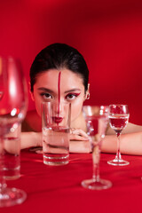 sensual asian woman with artistic makeup looking at camera near glasses with pure water on red background.