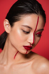 Fototapeta premium portrait of brunette asian woman with creative visage and naked shoulder posing isolated on red.