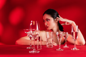 nude asian woman with creative makeup looking away near glasses with clear water on red background with shadow.
