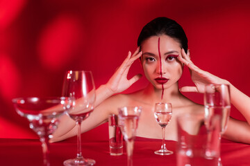 asian woman with bare shoulders and artistic makeup looking at camera near blurred glasses with water on red background.