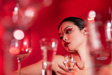 low angle view of nude asian woman with bright creative visage touching bare shoulder near blurred glasses on red background.