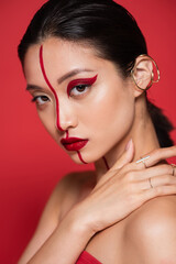 portrait of seductive asian woman with ear cuff and bright artistic visage touching bare shoulder isolated on red.