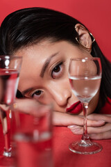portrait of young asian model with makeup looking at camera near blurred glasses with water isolated on red.